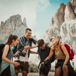 Group of smiling hikers with backpacks and cameras pose together on a rocky trail with towering mountain cliffs in the background.
