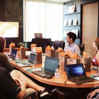 People seated around a conference table with laptops; a presenter stands at the front with a screen. Gift bags with orange tissue on the table.