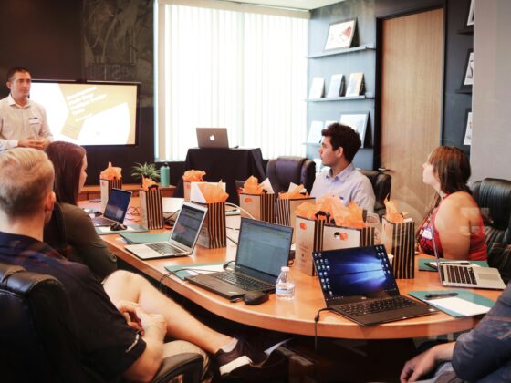 People seated around a conference table with laptops; a presenter stands at the front with a screen. Gift bags with orange tissue on the table.