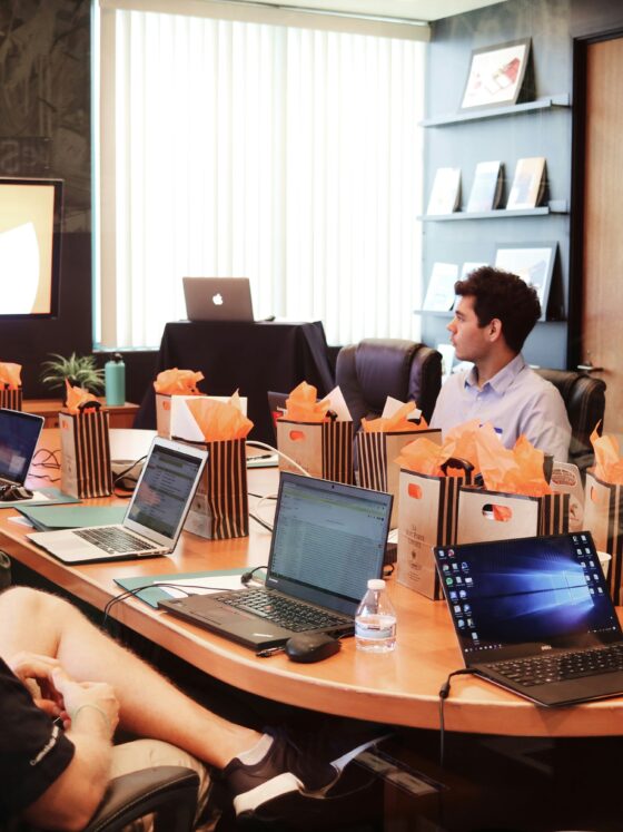 People seated around a conference table with laptops; a presenter stands at the front with a screen. Gift bags with orange tissue on the table.