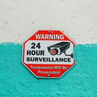 Red octagonal warning sign saying 'WARNING 24 HOUR SURVEILLANCE' with a camera icon, mounted on a teal-painted wall.