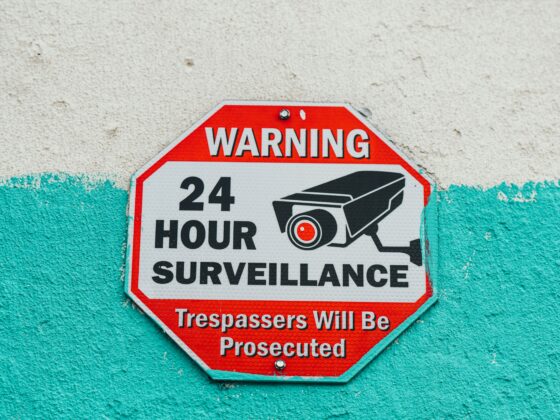 Red octagonal warning sign saying 'WARNING 24 HOUR SURVEILLANCE' with a camera icon, mounted on a teal-painted wall.
