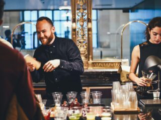 Bearded man in a dark shirt smiles while pouring a bottle at a busy bar counter with a woman preparing drinks in the background