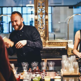 Bearded man in a dark shirt smiles while pouring a bottle at a busy bar counter with a woman preparing drinks in the background
