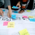 People collaborate around a table, sticking colorful notes on a large planning sheet filled with diagrams and comics.