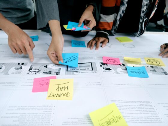 People collaborate around a table, sticking colorful notes on a large planning sheet filled with diagrams and comics.