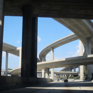 Complex freeway interchange with multiple elevated concrete ramps and pillars against a blue sky; cars visible on the lower roadway.