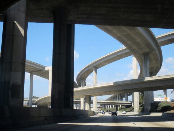 Complex freeway interchange with multiple elevated concrete ramps and pillars against a blue sky; cars visible on the lower roadway.