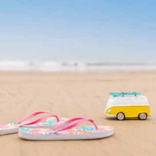 Toy yellow and white camper van with a turquoise surfboard on top sits on a sandy beach, with colorful flip-flops in the foreground.