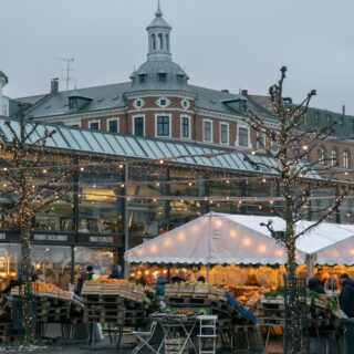 Winter market in a cobblestone square with string lights and produce stalls; a historic brick building with a domed tower in the background.