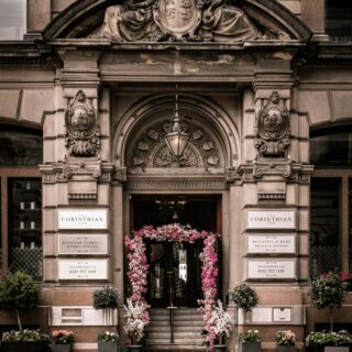 Ornate city building entrance with sculpted carvings and a pink flower arch framing the doorway, sign reads 'The Corinthian Club'