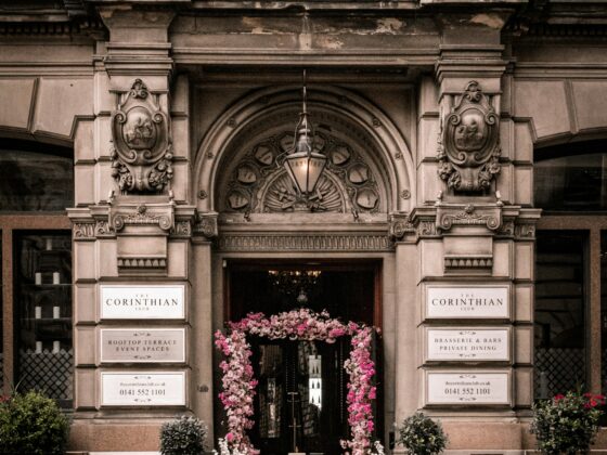 Ornate city building entrance with sculpted carvings and a pink flower arch framing the doorway, sign reads 'The Corinthian Club'