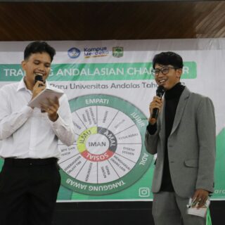 Two men on a stage with microphones; one reads from a notebook, the other smiles in a suit, behind them a wheel banner.