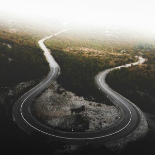 Aerial view of a winding mountain road curving through autumn-colored forest, with a hazy white sky above.