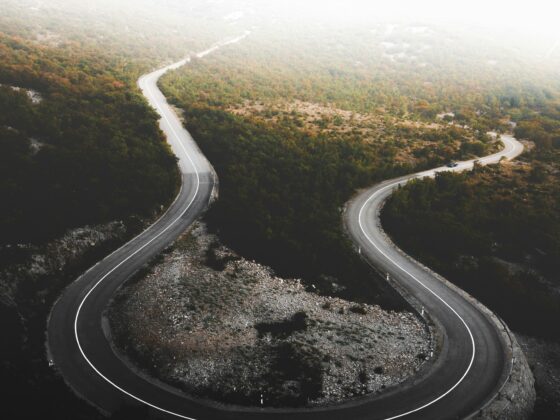 Aerial view of a winding mountain road curving through autumn-colored forest, with a hazy white sky above.