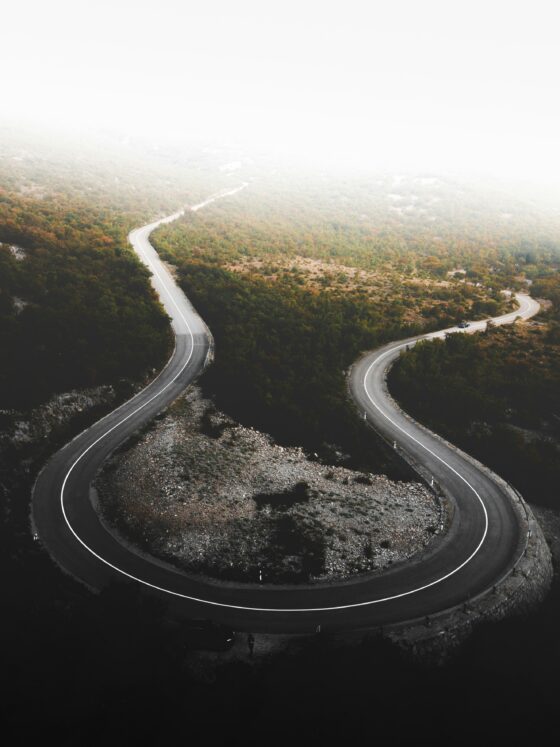 Aerial view of a winding mountain road curving through autumn-colored forest, with a hazy white sky above.
