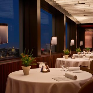 Elegant rooftop restaurant at night with round tables, white cloths, candles, potted plants, and city lights visible through windows.
