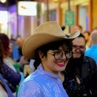 Smiling woman in a sparkly blue outfit wearing a tan cowboy hat and glasses at a crowded nightlife street.