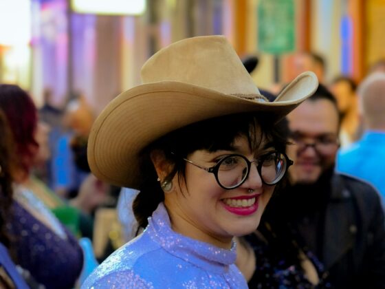 Smiling woman in a sparkly blue outfit wearing a tan cowboy hat and glasses at a crowded nightlife street.