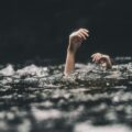 Hands raised above dark water, as if reaching for help, with a cloudy, stormy backdrop behind.