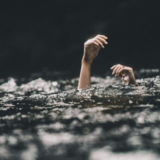 Hands raised above dark water, as if reaching for help, with a cloudy, stormy backdrop behind.