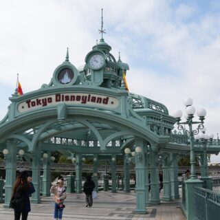 Ornate teal Tokyo Disneyland entrance with a round clock tower and a castle emblem; guests walk beneath on a cloudy day.
