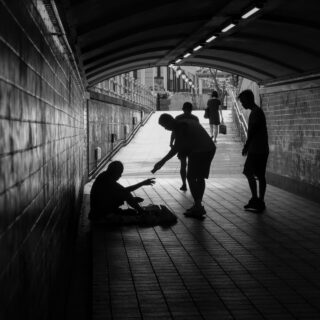 Silhouetted people in a long, arched pedestrian tunnel, some sitting on the floor and others standing as they approach the light at the exit.