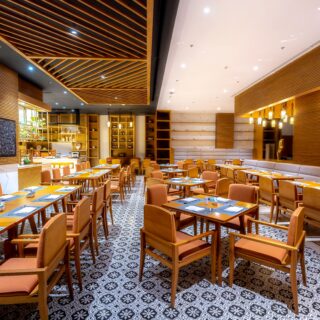 Bright, modern restaurant dining area with wooden tables and tan chairs, patterned tile floor, and warm pendant lighting.