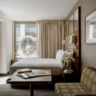 Hotel room with a king bed, beige curtains, a seating area, and a large window overlooking a decorative building façade.