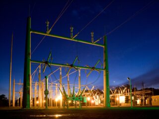 Nighttime view of a high-voltage electrical substation with green steel framework and insulators, illuminated by warm orange lights against a dark blue sky
