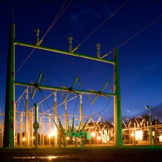 Nighttime view of a high-voltage electrical substation with green steel framework and insulators, illuminated by warm orange lights against a dark blue sky