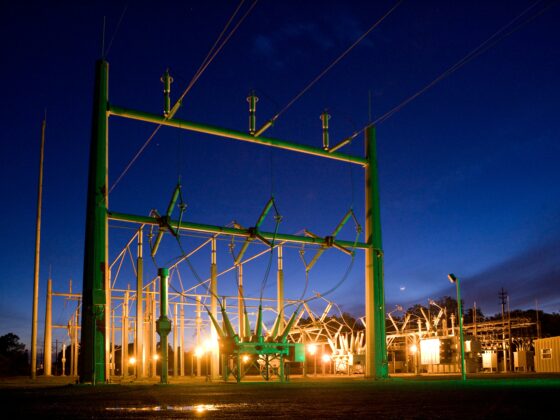 Nighttime view of a high-voltage electrical substation with green steel framework and insulators, illuminated by warm orange lights against a dark blue sky
