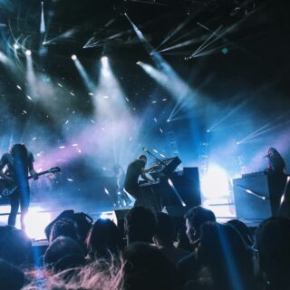 Band performs on a dark stage with blue and white spotlights, guitarist on left and keyboardist center, audience at front.