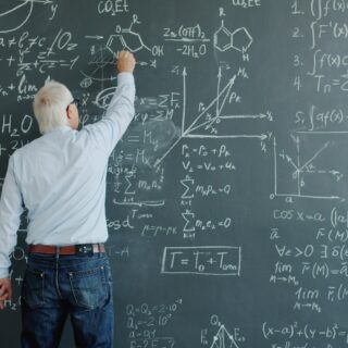 A person with white hair writing equations on a chalkboard filled with math formulas and diagrams, indicating a classroom teaching moment.