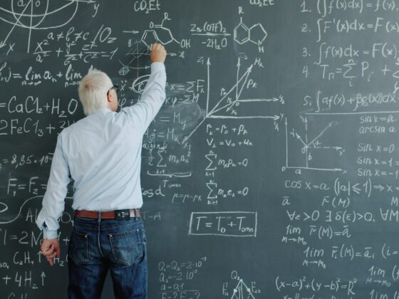 A person with white hair writing equations on a chalkboard filled with math formulas and diagrams, indicating a classroom teaching moment.