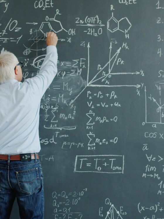 A person with white hair writing equations on a chalkboard filled with math formulas and diagrams, indicating a classroom teaching moment.