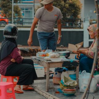 Three people gathered around a makeshift street-food table with bowls and containers, outdoors in a city square.