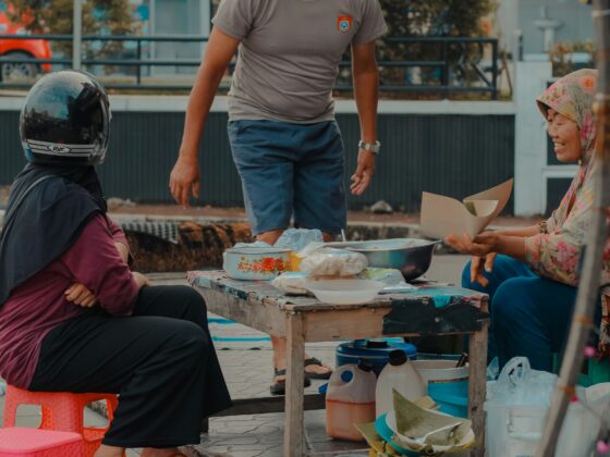 Three people gathered around a makeshift street-food table with bowls and containers, outdoors in a city square.