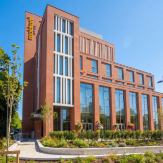 Brick, multi-story building with large glass windows and a landscaped front plaza under a clear blue sky, suggesting a hotel or office complex.