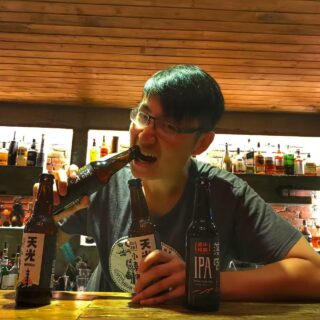 Man with glasses biting into a beer bottle at a bar, two bottles on the counter, backlit liquor shelves behind him nearby.
