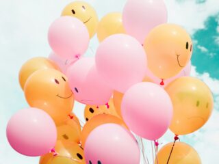 Cluster of pink and orange smiling balloons floating against a blue sky, tied with ribbons.