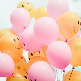 Cluster of pink and orange smiling balloons floating against a blue sky, tied with ribbons.