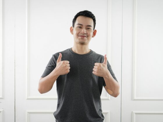 Smiling man in a gray T-shirt gives two thumbs up in front of a white paneled wall, showing a positive gesture.