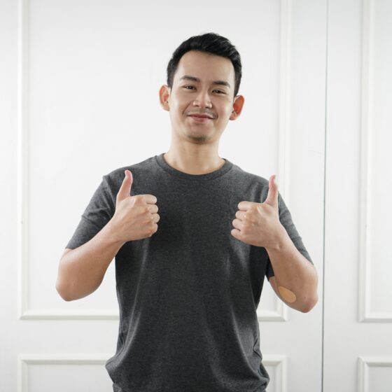 Smiling man in a gray T-shirt gives two thumbs up in front of a white paneled wall, showing a positive gesture.