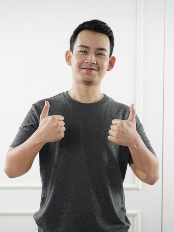 Smiling man in a gray T-shirt gives two thumbs up in front of a white paneled wall, showing a positive gesture.