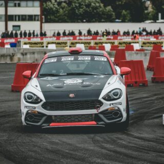 Race car navigating a course lined with red barriers and a crowd watching from behind barriers in a race venue.