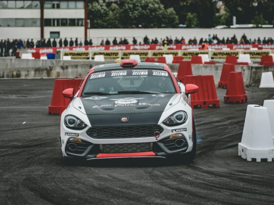 Race car navigating a course lined with red barriers and a crowd watching from behind barriers in a race venue.