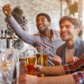 Three friends at a bar raise their glasses and cheer, smiling at the camera.”