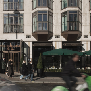 Urban street scene in front of The Athenaeum hotel with pedestrians and a green scooter passing by at street level.