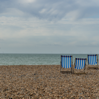 Four blue-and-white striped beach chairs on a pebbled shore facing the calm sea under a cloudy sky nearby.